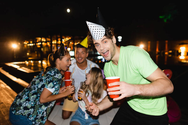 Selective focus of young man in party cap holding disposable cup near friends during party at night 