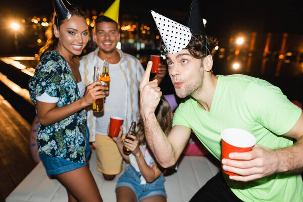 Selective focus of man with disposable cup pointing at party cap near friends with beer at night 