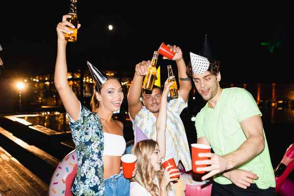 Young friends in party caps holding bottles of beer outdoors at night 