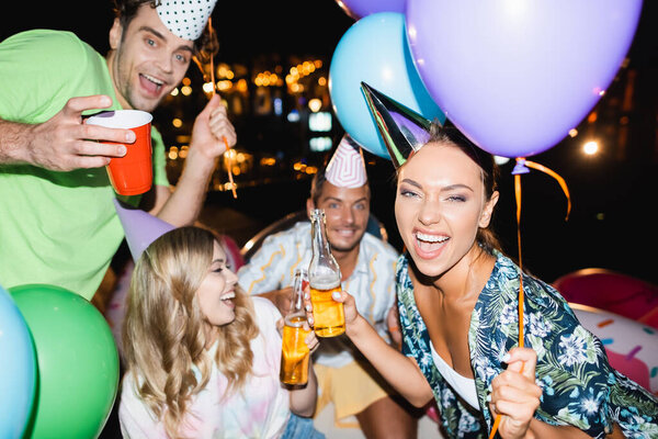 Selective focus of woman holding beer bottle and balloon during party with friends at night 