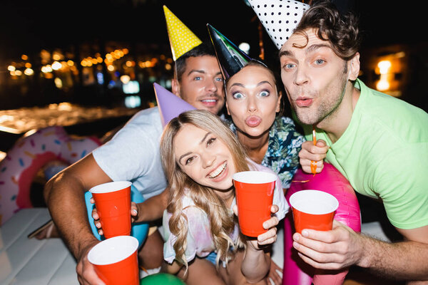Selective focus of couples in party caps looking at camera while holding disposable cups at night 