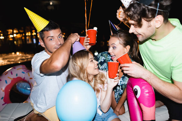 Selective focus of friends toasting with disposable cups near balloons during party at night 