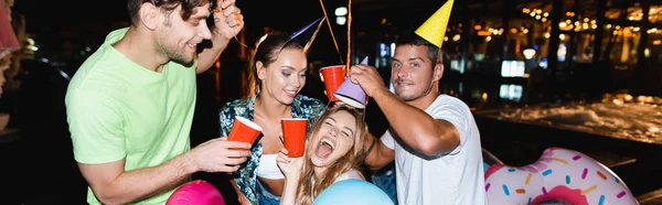 夜にプールの近くでパーティー中に使い捨てカップを保持する友人のパノラマショット — ストック写真