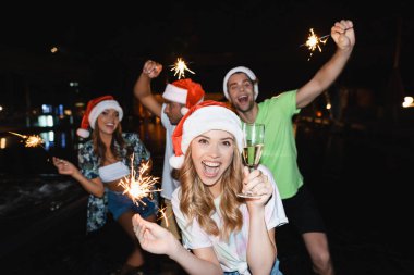 Seçici odak noktası Noel Baba şapkalı bir kadın elinde şampanya bardağı ve geceleri arkadaşlarının yanında maytap tutuyor. 