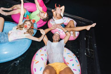 High angle view of young people in party headbands with blow ticklers swimming on rings in pool at night  clipart