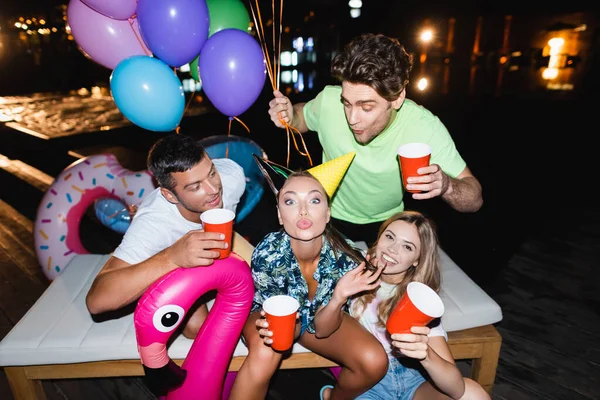 Young woman in party cap looking at camera near friends with disposable cups and balloons at night