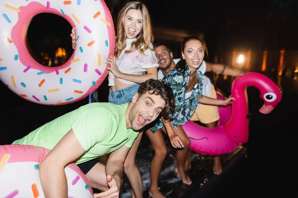 Selective focus of young man with swim ring looking at camera near friends at night