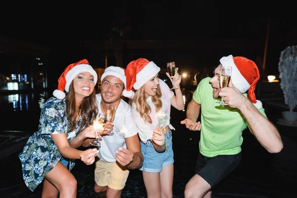 Vänner Santa Hattar Som Håller Tomtebloss Och Champagne Nära Poolen — Stockfoto