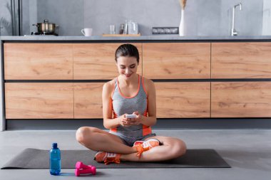Sporcu kadın, mutfaktaki su şişesi ve dambılların yanında fitness minderi üzerinde akıllı telefon kullanıyor. 