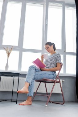 Pencere kenarında kitap okurken elinde bardak tutan çıplak ayaklı kadın. 