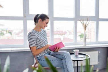 Akıllı telefon ve fincanın yanında kitap okuyan esmer kadının seçici odağı. 
