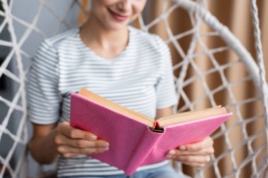 Evdeki sallanan koltukta kitap okuyan genç bir kadın görüntüsü. 