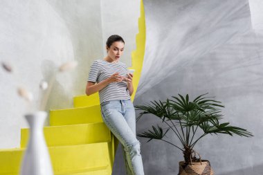 Şok olmuş bir kadının evdeki merdivenlerin yanında akıllı telefon kullanmasının seçici odağı 
