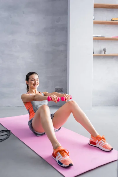 带哑铃的女运动员在家里的健身垫上做腹肌 — 图库照片