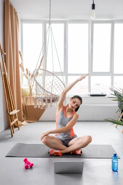 年轻女子在家里的笔记本电脑 水和哑铃旁边的健身垫上进行训练时伸懒腰 — 图库照片