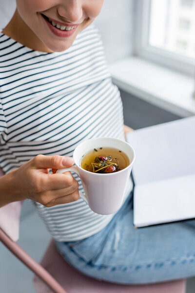 Cropped view of young woman holding cup of herbal tea and notebook at home 