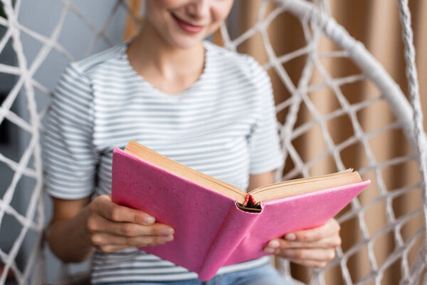 Cropped view of young woman reading book in hanging armchair at home 