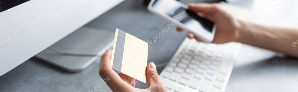 Panoramic shot of freelancer holding credit card and smartphone near computer 