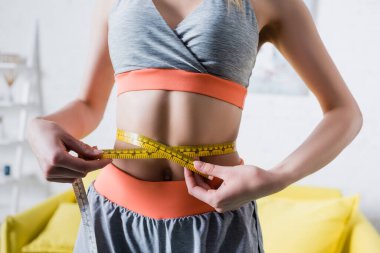 Cropped view of sportswoman measuring thin waist with tape at home clipart
