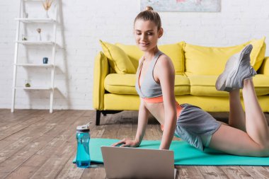 Spor kadını laptopun yanında egzersiz yapıyor ve evdeki spor minderinde bir şişe su içiyor.