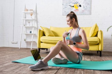 Gülümseyen sporcu kadın spor şişesini tutarken evdeki spor minderinde oturuyor. 
