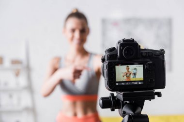 Dijital kameranın yanındaki spor şişesini gösteren gülümseyen sporcu kadının seçici odağı 
