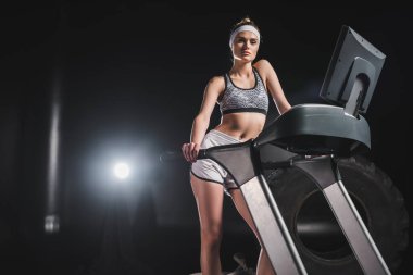 Sportswoman looking at camera while standing on treadmill in gym  clipart
