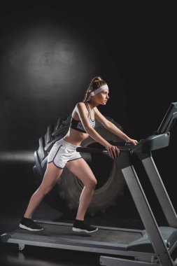 Sportswoman working out on treadmill near tire in gym  clipart