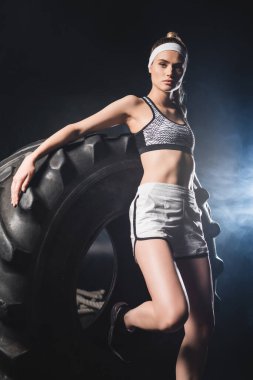 Spor giyim ve saç bandı giymiş genç bir kadının spor salonundaki lastik yakınında dumanla kameraya baktığı seçici odak noktası. 