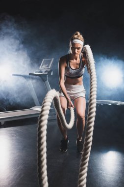 Spor salonunda dumanlı savaş halatıyla egzersiz yapan sporcu kadınların seçici odağı. 