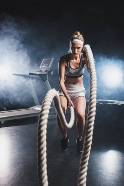 Selective focus of sportswoman exercising with battle rope in sports center with smoke  clipart