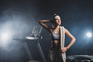 Savaş halatıyla genç sporcu kadın lastik ve koşu bandının yanından dumanla uzağa bakıyor. 