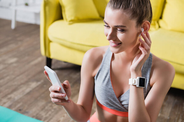 Sportswoman with wireless earphone and smartwatch holding cellphone at home