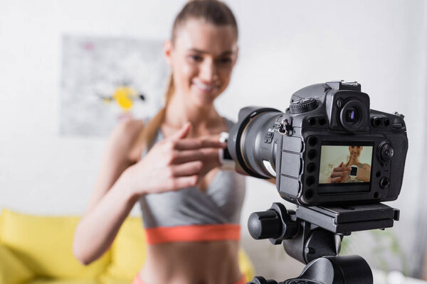 Выборочный фокус улыбающейся спортсменки, указывающей на умные часы возле цифровой камеры 