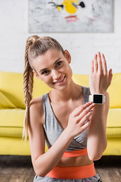 Smiling sportswoman touching fitness tracker and looking at camera at home