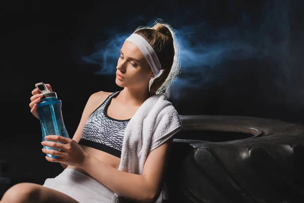 煙とジムでタイヤの近くのスポーツボトルを見てタオルと若いスポーツ女性の選択的な焦点 — ストック写真
