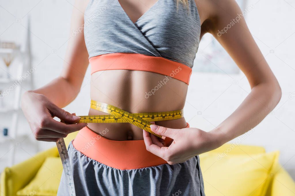 Cropped view of sportswoman measuring thin waist with tape at home