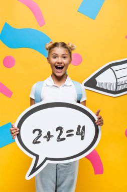 Heyecanlı okul çocuğu elinde matematik illüstrasyonuyla kağıt elementler ve sarı kalemlerin yanında konuşma balonu tutuyor. 