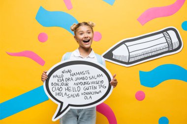 excited schoolchild holding speech bubble with greeting lettering near multicolored paper elements and pencil on yellow  clipart