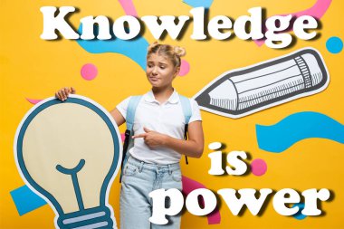 Confused schoolgirl pointing with finger at paper light bulb near decorative elements and knowledge is power lettering on yellow  clipart