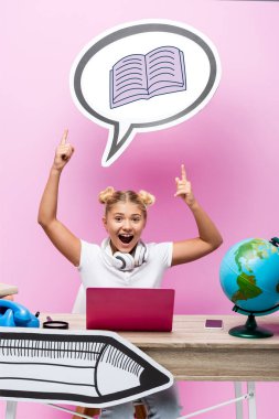 Heyecanlı çocuk parmaklarıyla konuşma balonunu işaret ediyor dizüstü bilgisayarının yanında kitap illüstrasyonuyla, akıllı telefonuyla ve pembe küreyle.