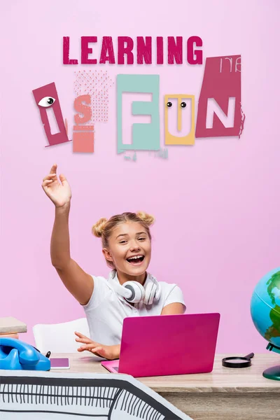 Schoolgirl sitting with raised hand near laptop, telephone, learning is fun lettering and paper art on pink