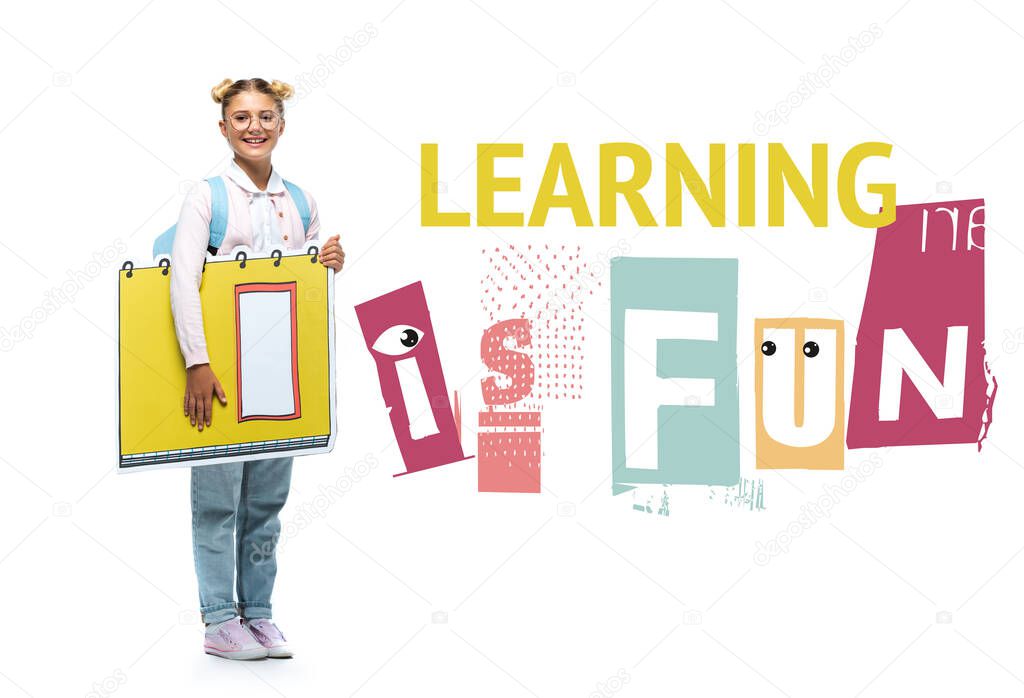 full length view of joyful schoolchild in eyeglasses holding paper cut notebook near learning is fun lettering on white