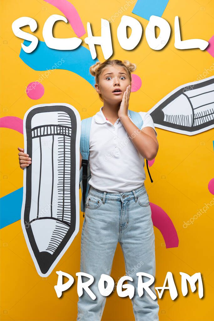 Worried schoolchild with backpack and paper pencil looking at camera near school program lettering on yellow 