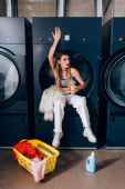 stylische Frau sitzt in der Waschmaschine neben Kunstpelzjacke und hält Orangensaft neben Korb mit schmutziger Kleidung im Waschsalon