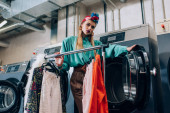 junge und stylische Frau im Turban steht neben Kleiderständern und Waschmaschinen im modernen Waschsalon 