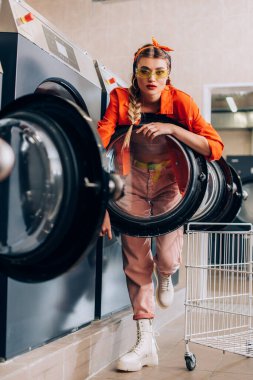 selective focus of stylish woman in sunglasses looking at camera near washing machines in laundromat clipart
