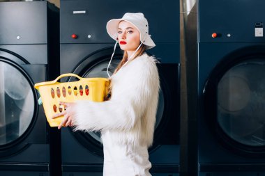 Sahte kürk ceketli şık bir kadın ve çamaşırhanedeki çamaşır makinelerinin yanında sarı sepetli bir şapka taşıyor.