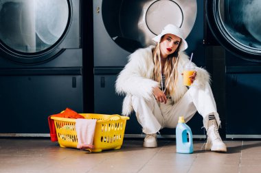 Sahte kürk ceketli şık bir kadın ve çamaşır makinesinde çamaşır ve çamaşır makineleriyle portakal suyuyla plastik bardak tutan bir şapka.