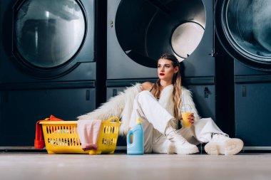 Sahte kürk ceketli şık bir kadın elinde portakal suyuyla çamaşır sepetinin yanında plastik bardakla çamaşır makinesinde deterjan şişesi ve çamaşır makinesi tutuyor.